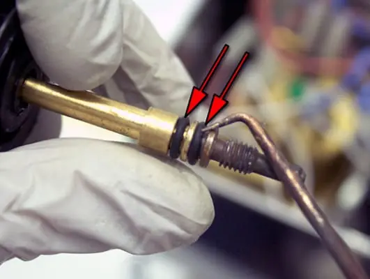 A gloved hand holds a brass shaft with two black O-rings—part of a Lelit PL41 Anna steam valve repair—highlighted by red arrows, next to a metal clamp or tool. 2025 1st-line.com