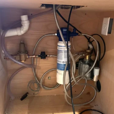 A water filtration system with hoses, pipes, and a filter cartridge is installed under a wooden sink cabinet. An electrical outlet, wiring, and plumbing fixtures are also visible. 2025 1st-line.com