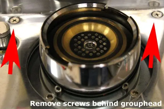 Close-up of a coffee machine grouphead with two red arrows pointing to screws behind it. White text at the bottom reads: Remove screws behind grouphead before removing top cover. 2025 1st-line.com