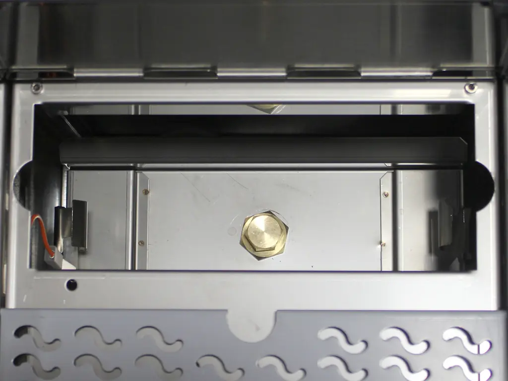 A close-up view of a metal appliance’s interior, possibly a grill or oven, featuring a hexagonal brass fitting—ideal for Strega switchable or Tank to Direct Connect Conversion setups—with cutout metal panels and a wavy-patterned bottom. 2025 1st-line.com