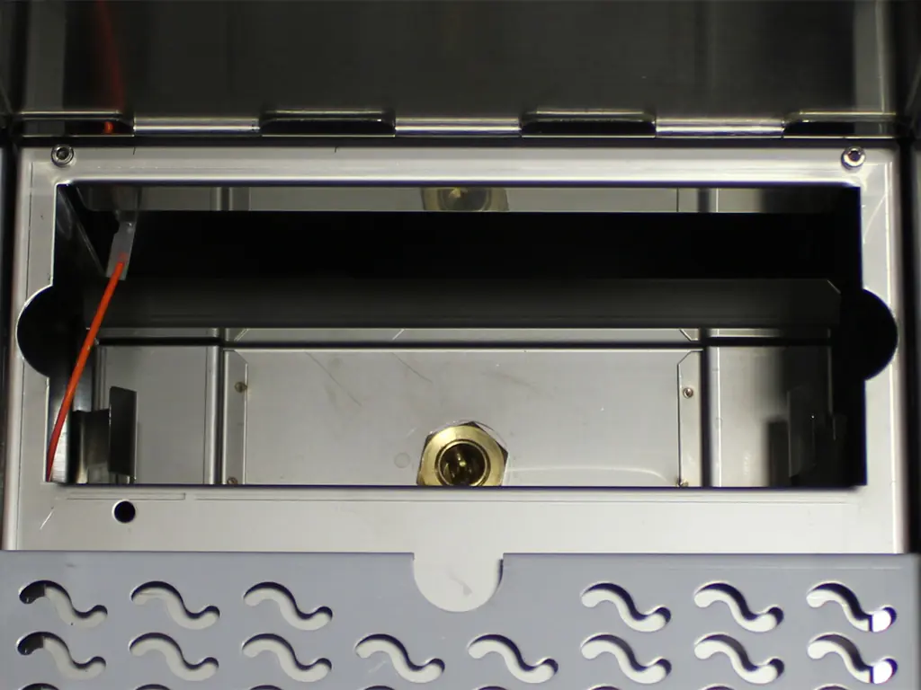 Close-up view of the inside of a stainless steel empty grill burner compartment, showing a rectangular opening with a gas inlet fitting and metal grate with wave-shaped cutouts—ideal for Tank to Direct Connect Conversion setups. 2025 1st-line.com