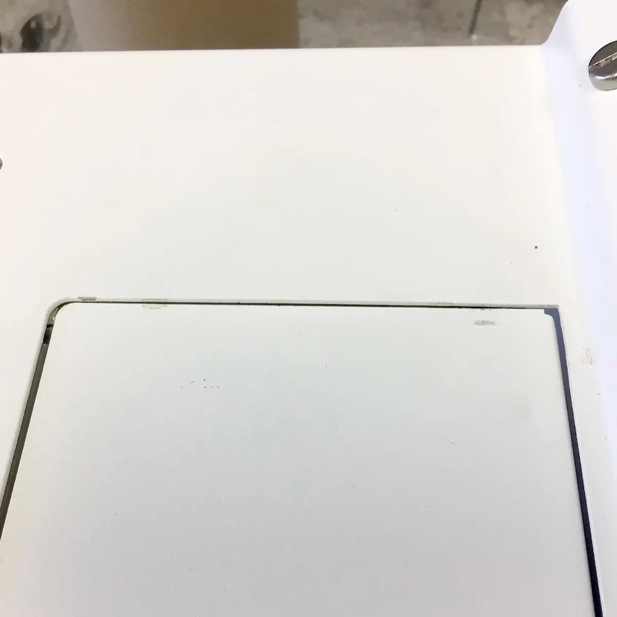 A close-up of a white rectangular panel with a faint outline and slight gap around the edge, set on the SIDEWALK SALE Nurri R-TYPE SB (HX) Espresso Machine (RETURNED & REFURBISHED); a metal screw is visible in the top right corner. 2025 1st-line.com