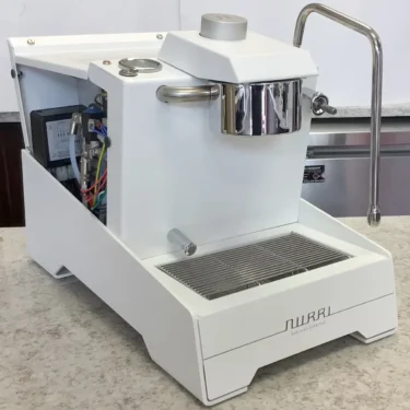 A refurbished Nurri R-TYPE SB (HX) espresso machine from the Sidewalk Sale, in modern white with a minimalist design, sits on a countertop. Internal components are visible on the left, and it includes a metal steam wand and drip tray. 2025 1st-line.com