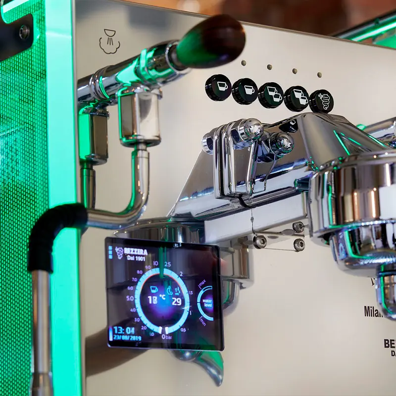 A close-up of a shiny espresso machine with a digital display showing temperature and time. The machine features metal dials, a portafilter, and a green LED light. Circular buttons for different drink options are visible above.
