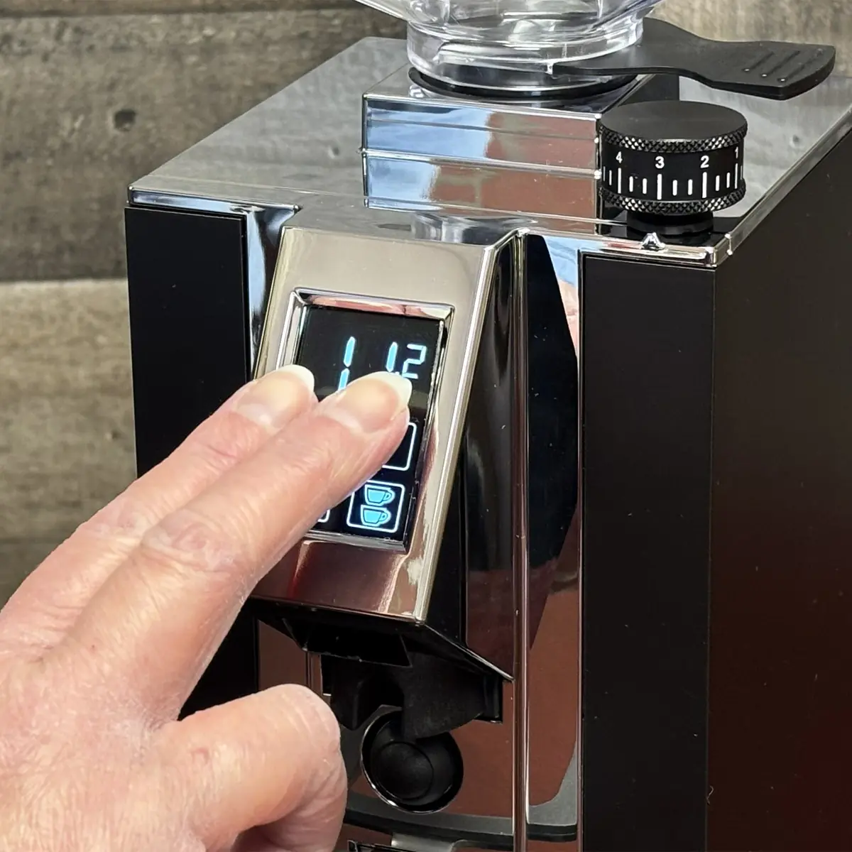A hand with two fingers is pressing buttons on the digital display panel of an Eureka Grinder coffee machine, showing the number 1.12. A grind size adjustment dial is visible on top.