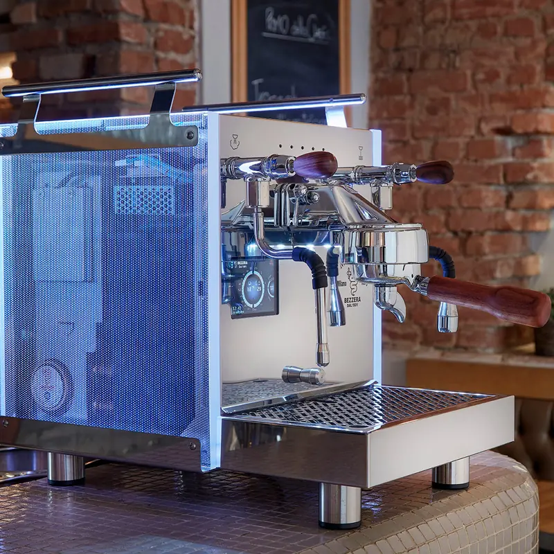 A modern espresso machine with a transparent side panel and wooden handles sits on a countertop in a café with exposed brick walls.
