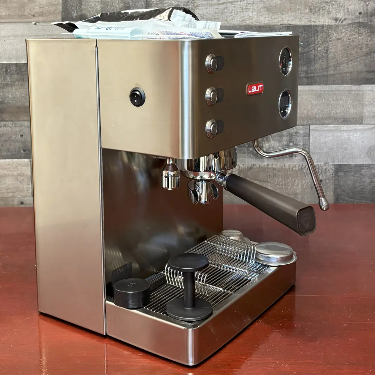A stainless steel espresso machine sits on a wooden surface with a steam wand, filter holder, and controls visible. Coffee-making accessories are placed on its drip tray. The background consists of wooden panels.