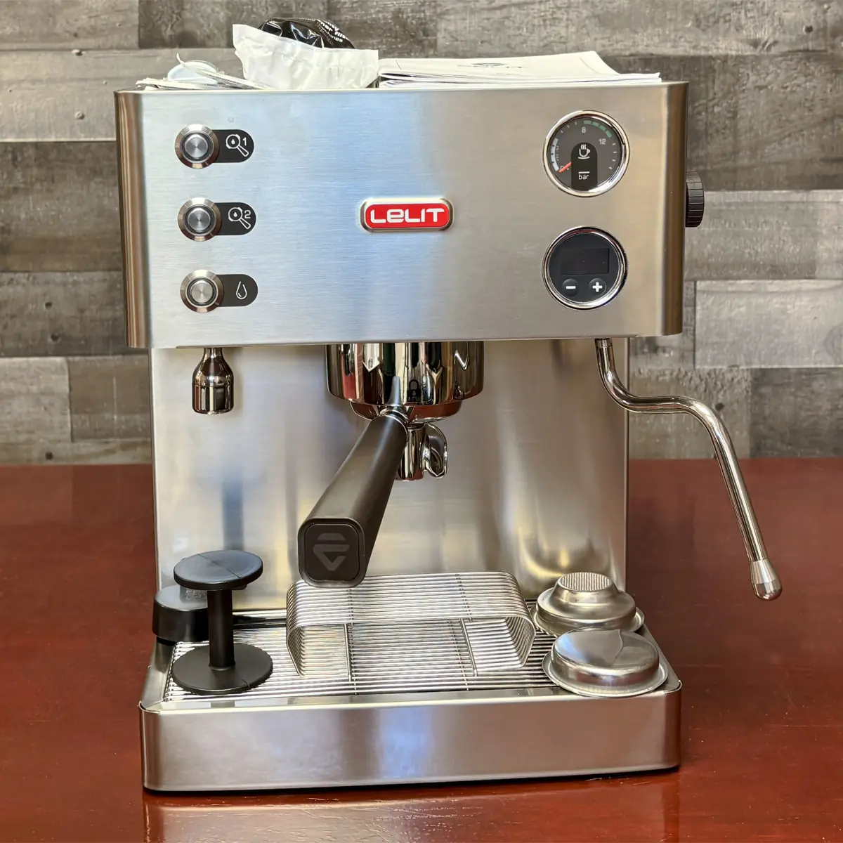 A stainless steel Lelit espresso machine sits on a wooden counter, featuring three buttons, a steam wand, a pressure gauge, portafilter, and tamper, with a rustic wooden wall in the background.