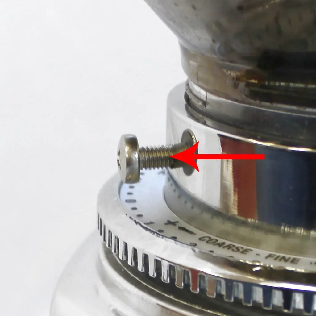 Close-up of a metal screw partially inserted into the shiny chrome surface of a Quamar Q50 coffee grinder, with a red arrow pointing to the screw. The background is white and part of a rotating dial with COARSE and FINE burrs settings is visible.