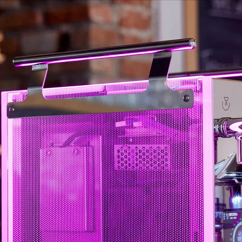 Close-up of a pink-lit metal coffee tamping station with a tamping mat and handle holder attached, against a blurred background with exposed brick wall.
