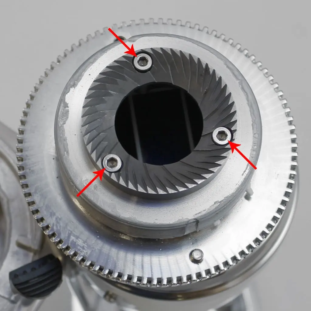 Close-up of a Quamar Q50 coffee grinder burr, with three red arrows highlighting screws that secure the burr to the grinder. The outer edge features fine, gear-like teeth characteristic of high-quality burrs.