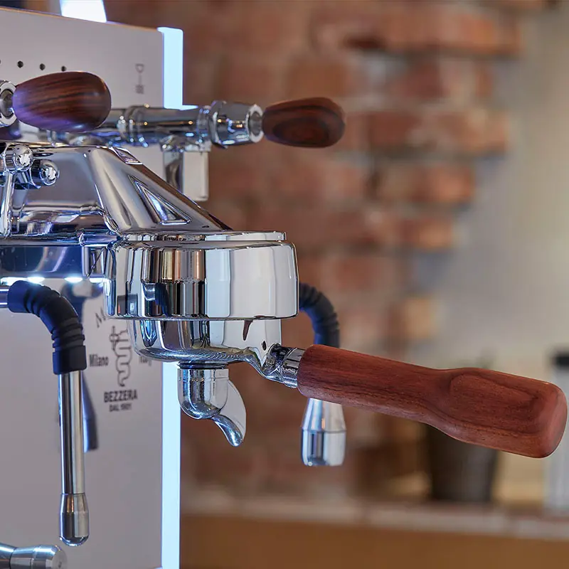 Close-up of a shiny espresso machine with wooden handles in a cozy indoor setting, featuring a blurred brick wall background.