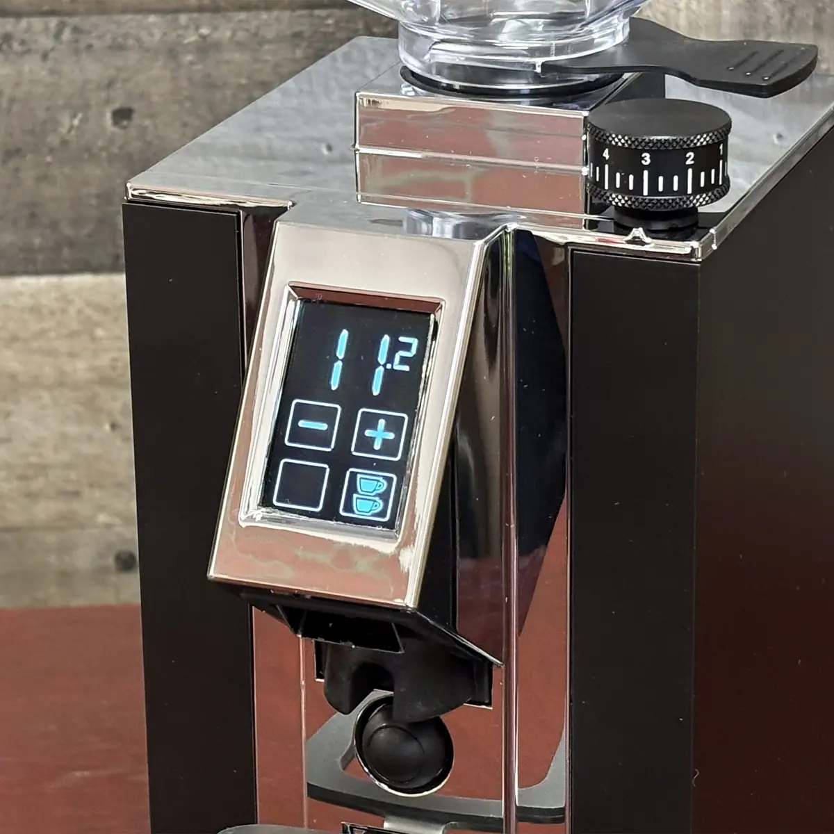 Close-up of a shiny, modern Eureka Grinder with a digital display showing 11.2 and touch buttons for settings. A black grind size dial sits on top, and the rustic wooden background highlights the sleek design—no missing buttons in sight.