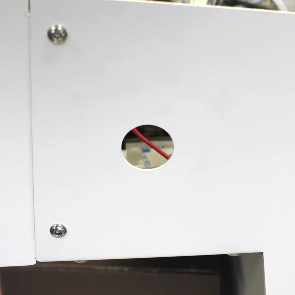Close-up of a white metal surface with a circular hole in the center, revealing red wires and internal components inside. Four screws secure the metal panels at the corners.