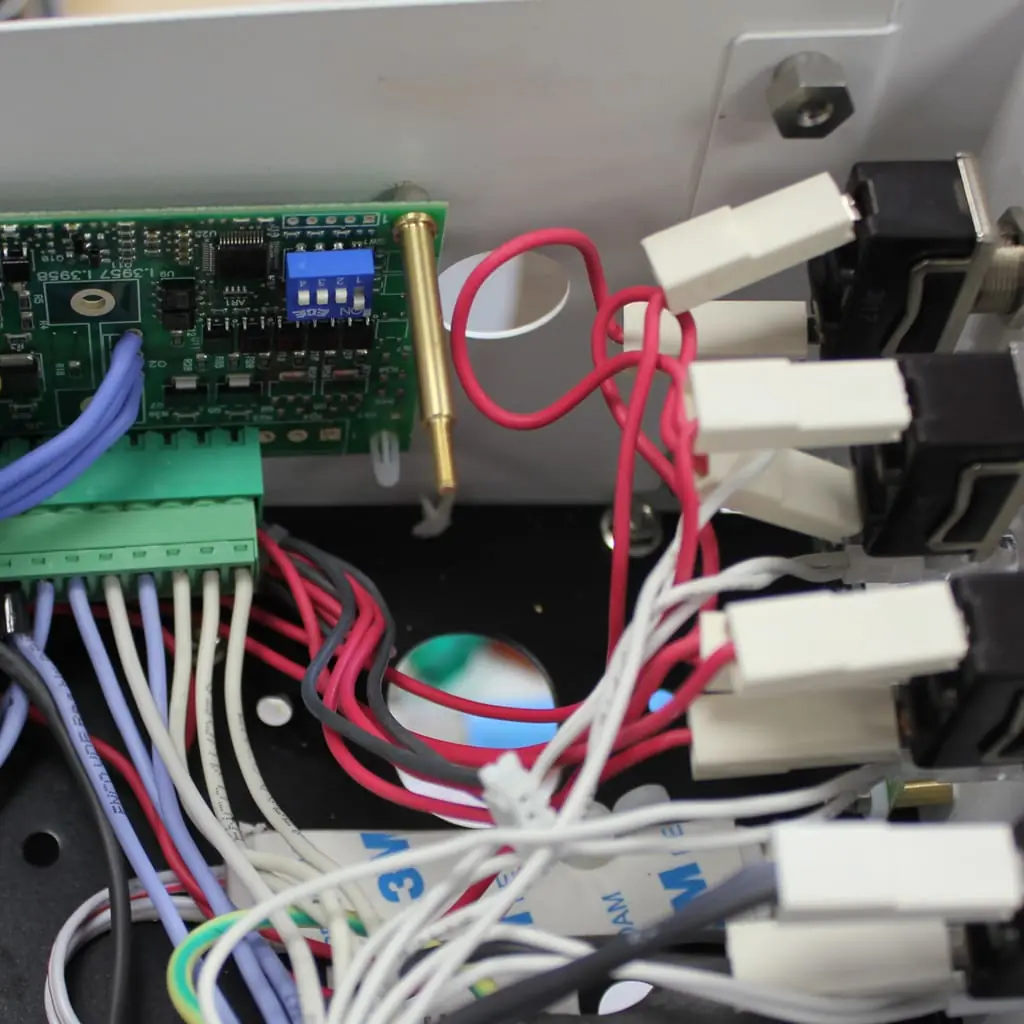 Close-up of an electronic circuit board with various colored wires connected to terminals and connectors inside a metal enclosure. Some wires are red, white, blue, and gray, and a green component is visible.