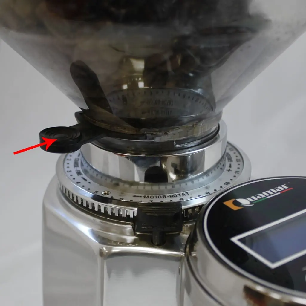 Close-up of the Quamar Q50 coffee grinder with a transparent bean hopper. A red arrow points to the adjustment knob on the collar, used to set burrs for precise grind size. The grinder features a shiny metallic finish and a digital display.