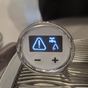 Close-up of an espresso machine display showing a warning symbol, a water tap icon, and a water droplet, with plus and minus buttons below. LELIT brand logo is visible at the bottom. 2025 1st-line.com