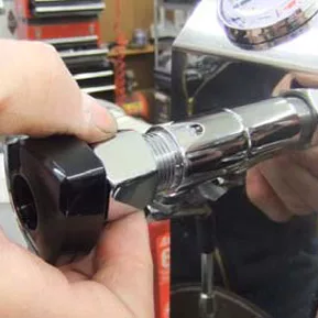 A person uses their hands to install or adjust a metal component with a black plastic knob on a machine, possibly performing hot water valve gasket repair. A gauge is visible in the background.