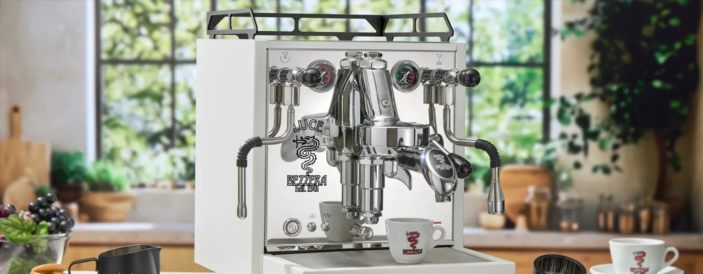 A Bezzera Luce HX Espresso Machine in white, featuring gauges and dual steam wands, sits on a kitchen counter. Two cups with a red logo are beneath the spouts as sunlight pours through large windows in the background.