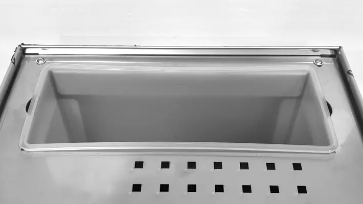 A rectangular metal container with a clear plastic lid fits in a recessed compartment of the Bezzera Luce HX Espresso Machine. Below, two rows of square ventilation holes are visible against a plain, light-colored background.