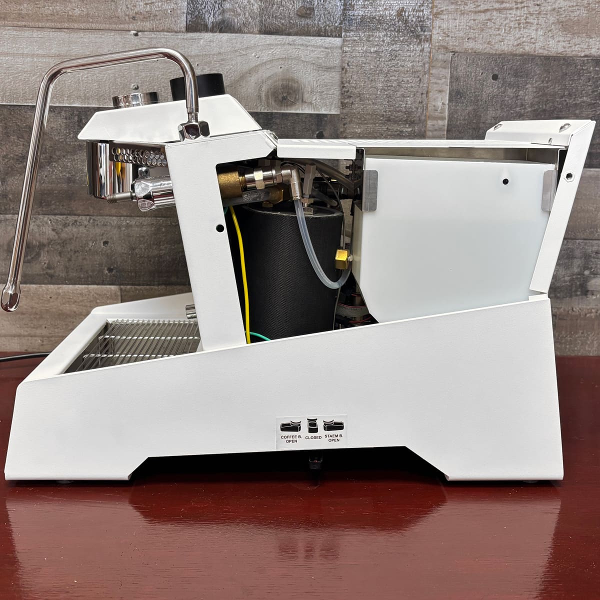 A white Nurri R-Type DB Dual Boiler Espresso Machine with its side panel removed shows internal components on a reddish-brown surface, set against a wooden wall.