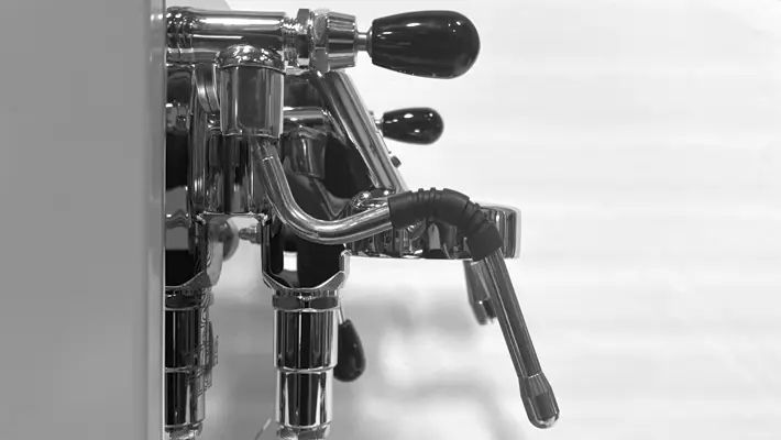 Close-up of the Bezzera Luce HX Espresso Machine’s sleek metal steam wand and portafilter handles, shot in black and white with a blurred background.