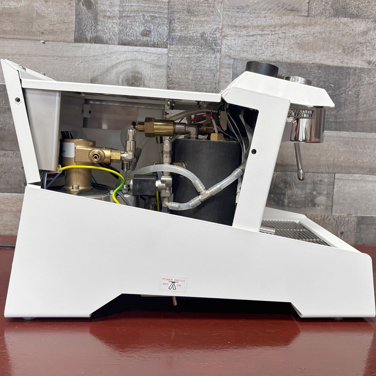 The Nurri R-Type DB Dual Boiler Espresso Machine, in white with its side panel removed to show internal pipes, wires, and the boiler, sits on a wooden surface against a wood-patterned wall.