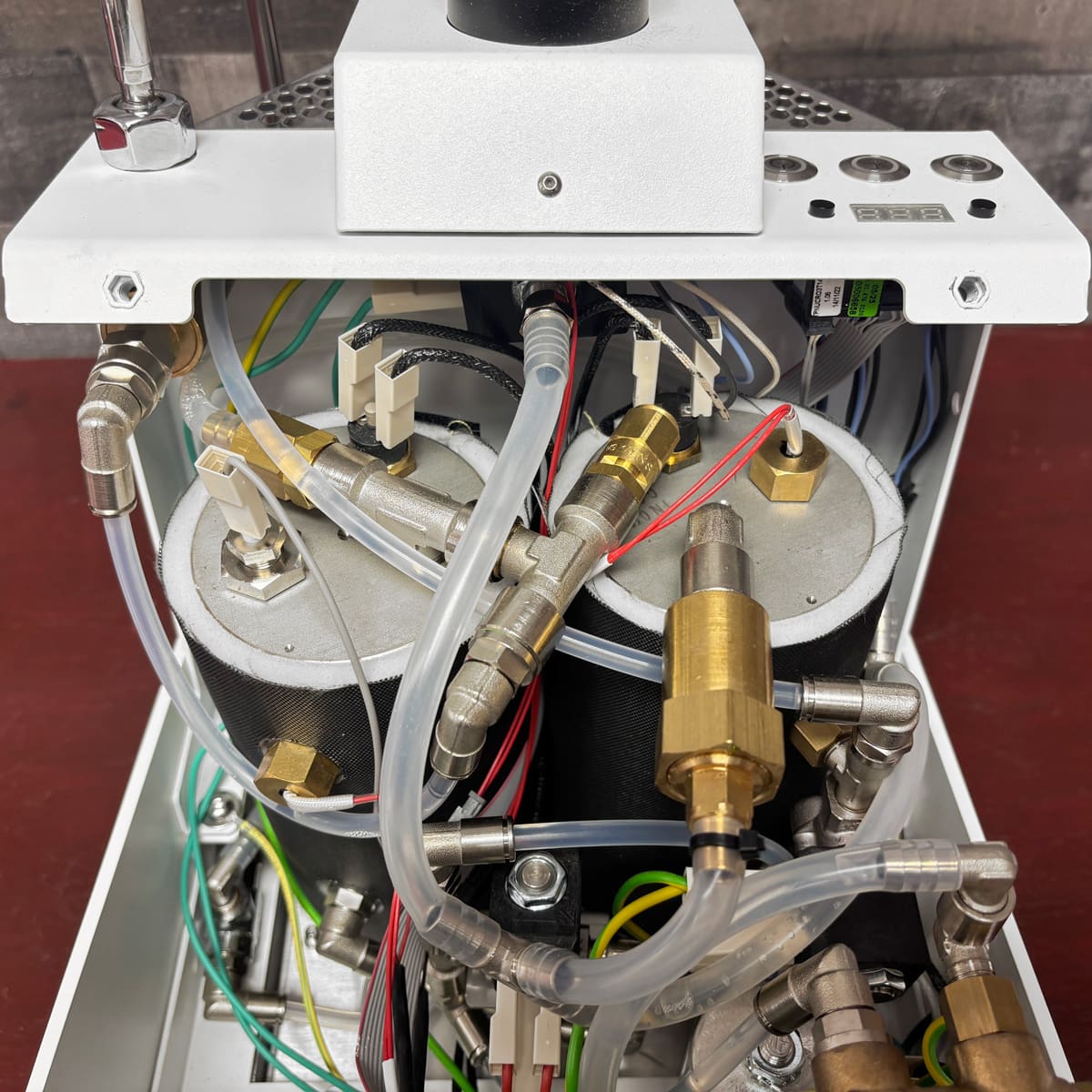 The Nurri R-Type DB Dual Boiler Espresso Machine is shown open, revealing two insulated cylindrical boilers, metal pipes, brass fittings, plastic tubes, wires, and connectors on a red surface.
