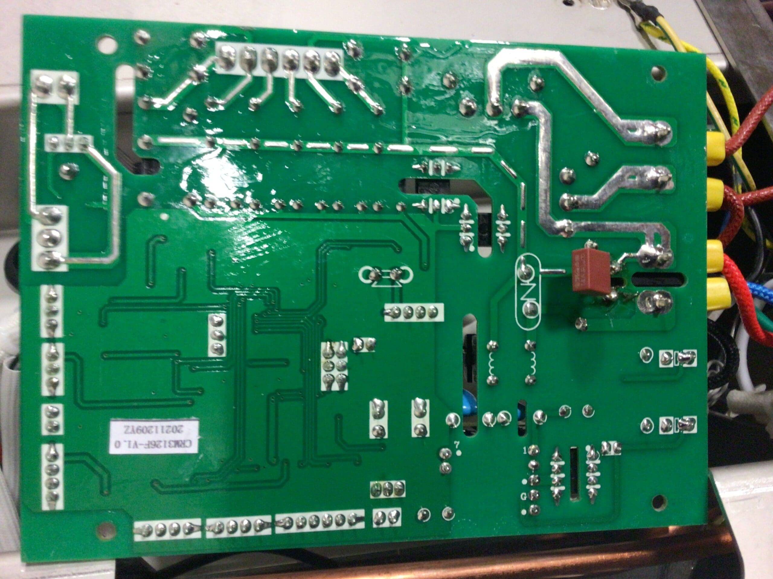 A close-up view of the underside of a green printed circuit board (PCB) shows various soldered connections, circuit traces, and several electronic components—including a fuse soldered to the Inizio Motherboard. Wires are seen connected at the edge.