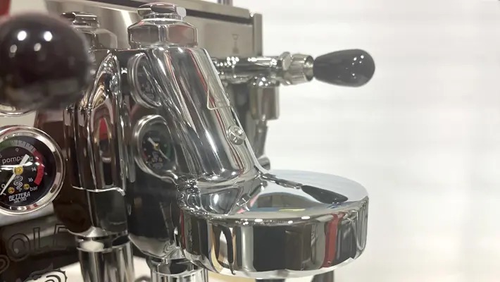 Close-up of a shiny chrome espresso machine group head with a pressure gauge, black lever, and various reflective metal components against a blurred white background.