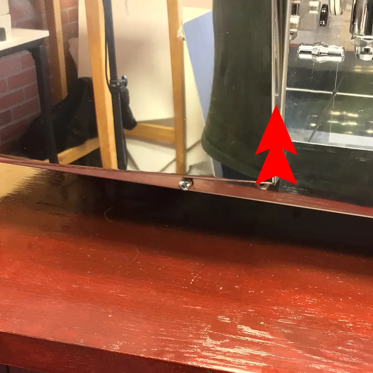 A red arrow points to a silver screw on the edge of a polished wooden surface, suggesting Accessing the Inside, with a mirror reflecting part of a person and some equipment in the background.
