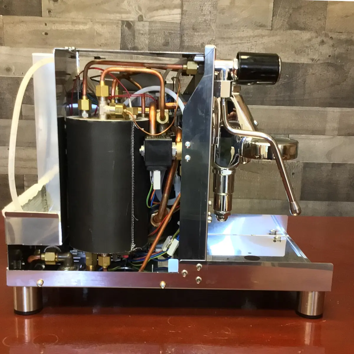 A Magister Stella semi-automatic espresso machine with its side panel removed reveals the inside—pipes, wiring, and a boiler—set on a red surface against a wood-patterned wall.