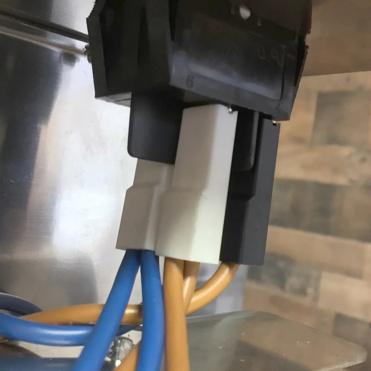 Close-up of electrical connectors with blue and brown wires attached, plugged into a black socket—offering a glimpse of accessing the inside—set against a metallic and wooden floor background.