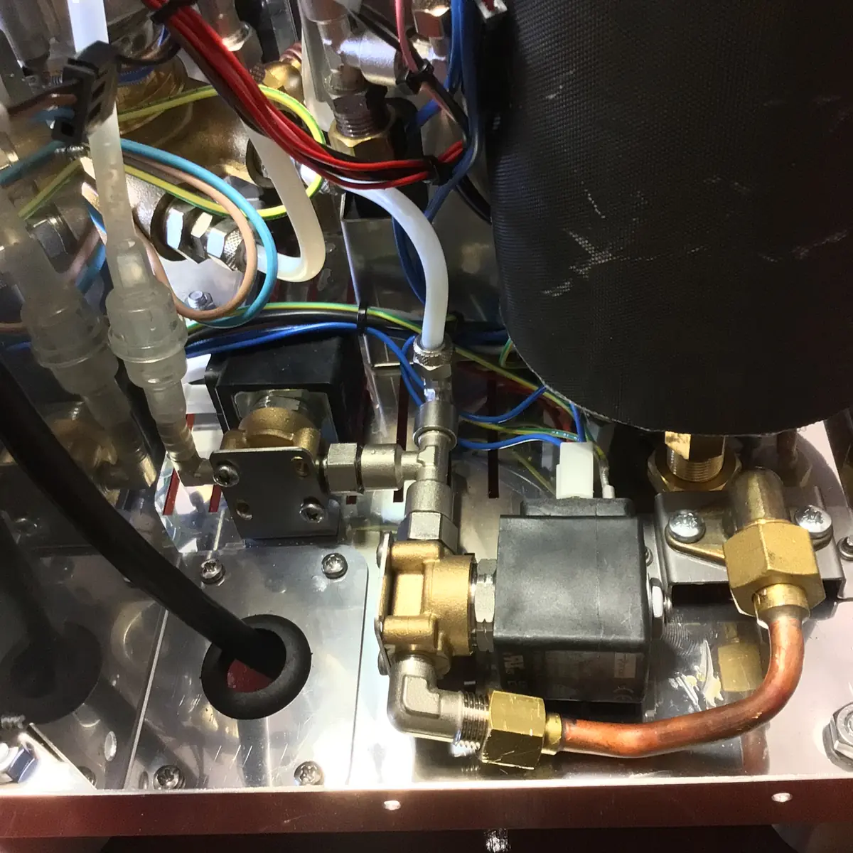 Close-up view inside a Magister Stella espresso machine, showcasing colored wires, metal pipes, valves, and connectors neatly arranged on a reflective metal base.