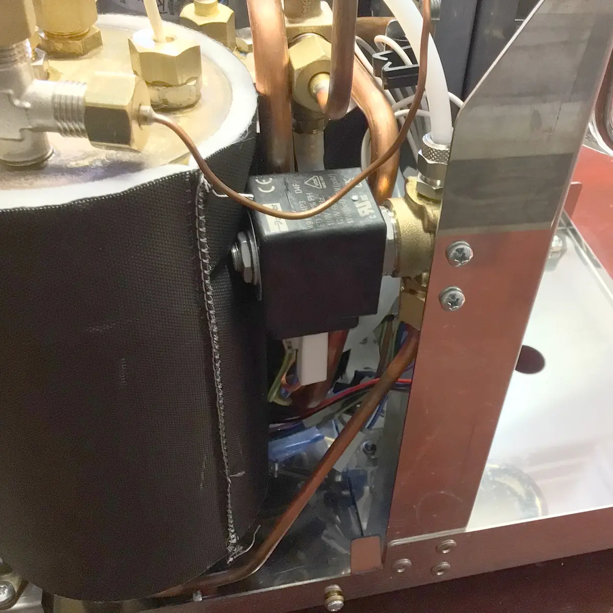 Accessing the Inside of a Magister Stella coffee machine reveals a close-up of its internal components: a cylindrical black-insulated boiler, copper pipes, brass fittings, electrical wiring, and a solenoid valve.