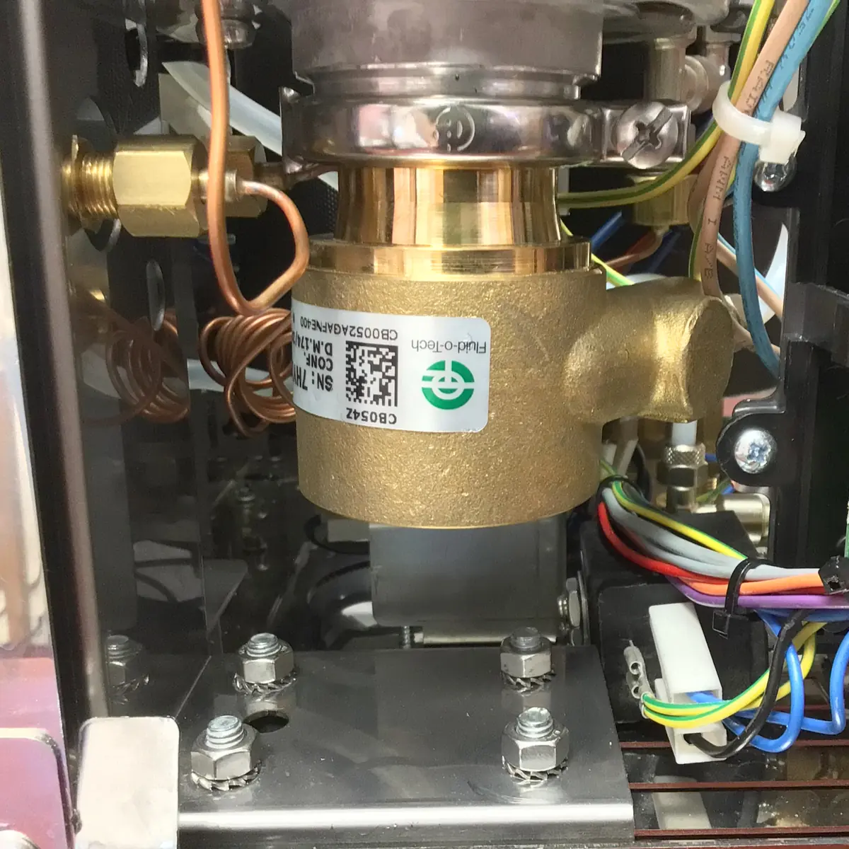 Close-up of internal components of a Magister Stella machine, showing metallic valves, pipes, copper coils, bolts, and multicolored wiring. A QR code and label are visible on a large brass valve at the center, accessing the inside.