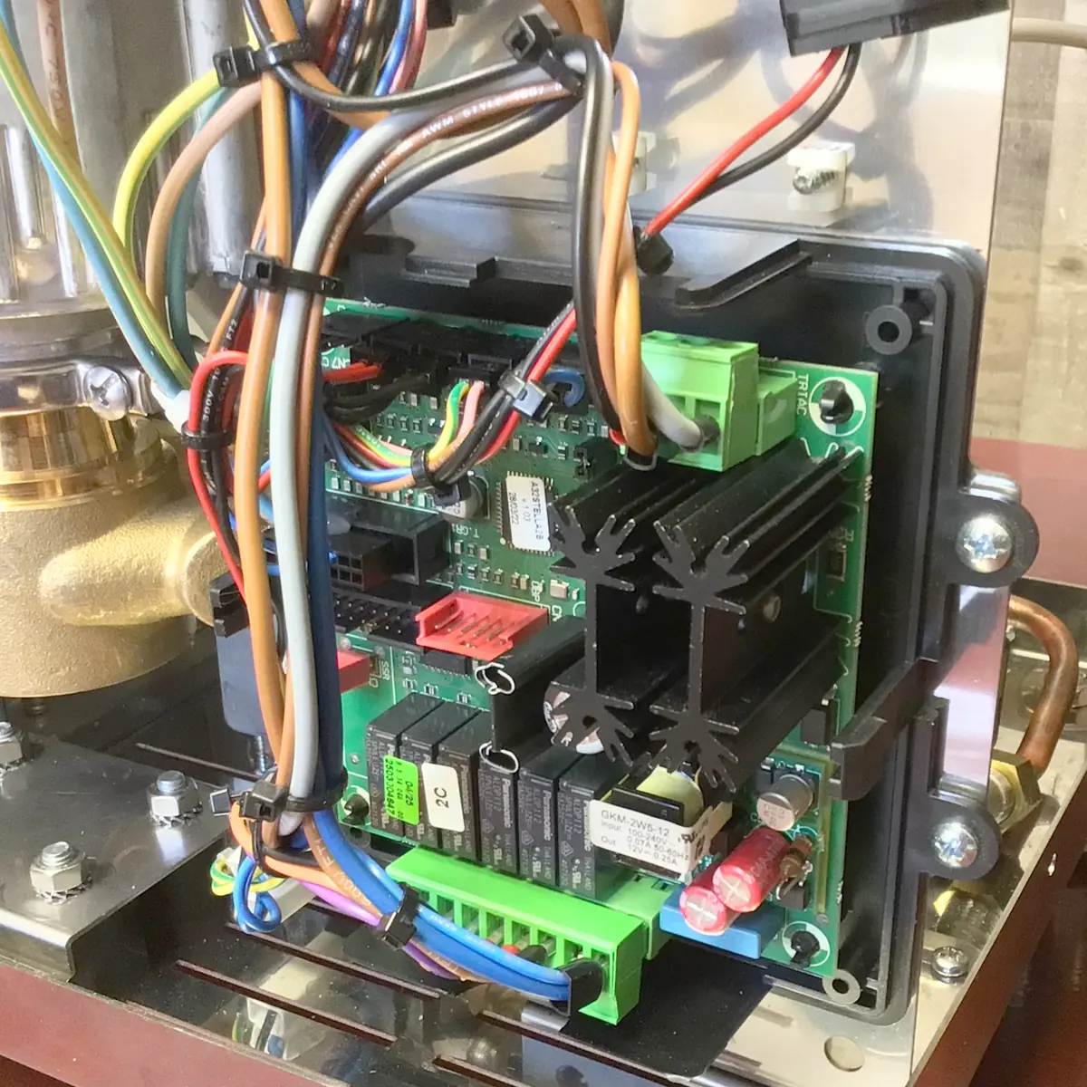 Close-up of an electronic circuit board with multicolored wires connected to various terminals and components, mounted inside a metallic Magister Stella enclosure. Black heat sinks and green connectors are visible on the board.