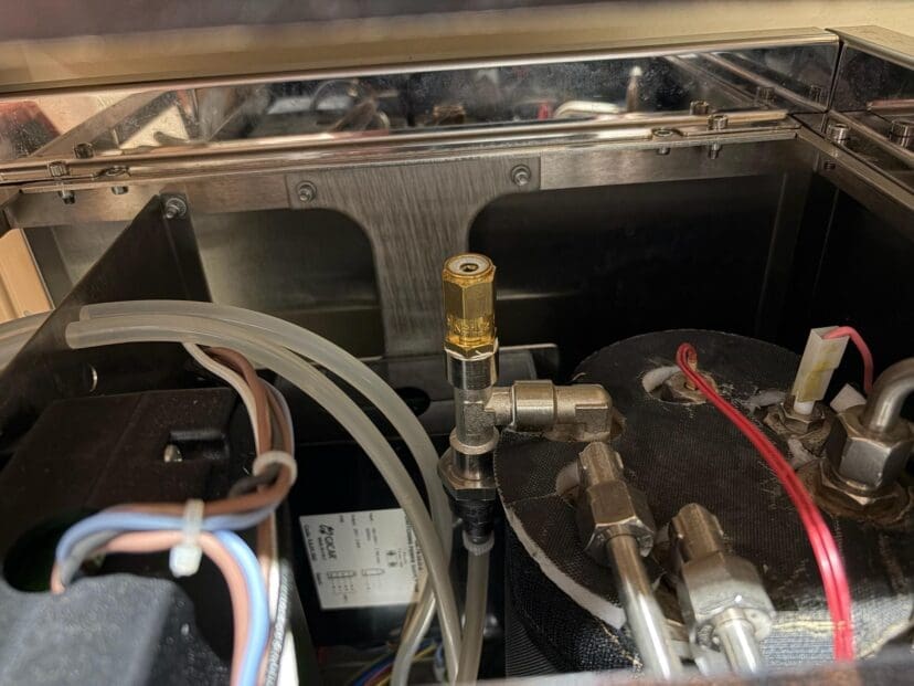 Close-up view of the internal components of a machine, showing various tubes, wires, metal fittings, and a brass valve in the center. The setup includes covered parts and connectors.