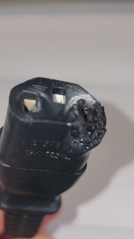 Close-up of a damaged black electrical plug with visible burn marks and melted plastic on one side, indicating it has overheated or experienced electrical damage.