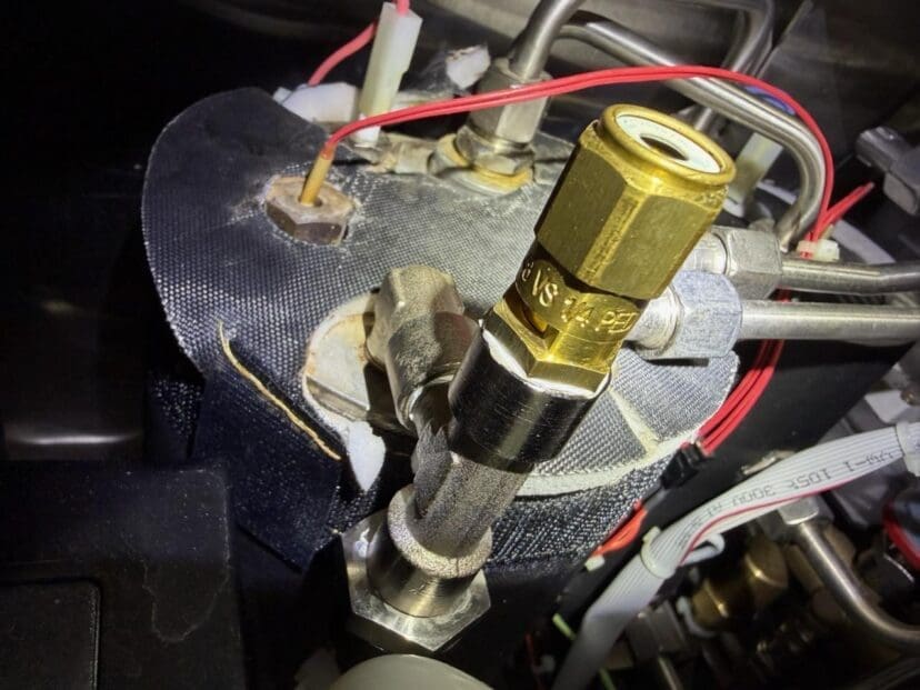 Close-up of a brass valve connected to metal pipes, surrounded by red and white wires, attached to a cylindrical device covered with dark insulating material.